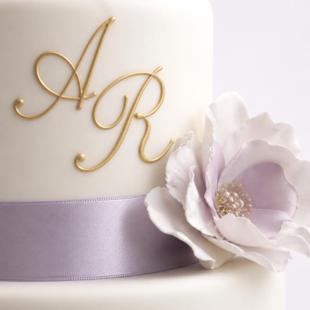 Close-up of a wedding cake showing a hand-piped gold monogram, a silk ribbon and a single delicate sugar flower